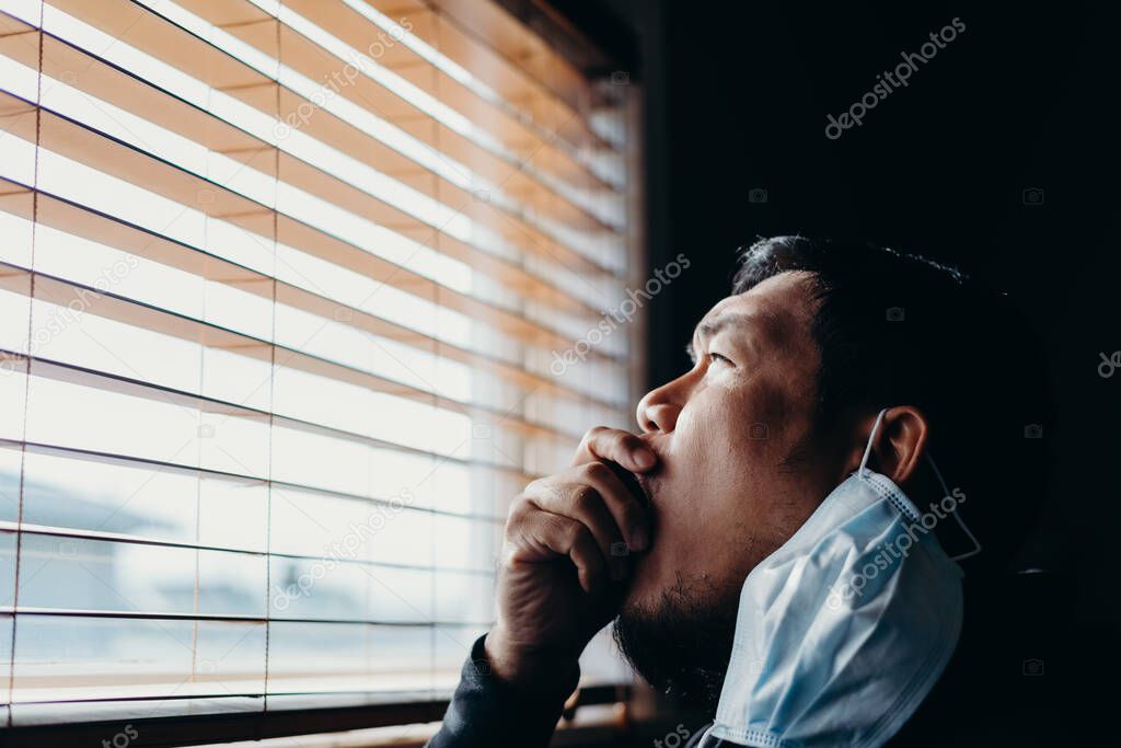 Asian man in isolation looking through window blinds and cough. Man in medical mask forced to stay inside the house as a result of the restrictions caused by the Coronavirus outbreak.