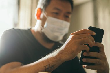 Koronavirüs karantinası kavramı. Tıbbi maskeli bir adam evde haber aramak için telefonu kullanıyor..