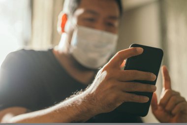 Koronavirüs karantinası kavramı. Tıbbi maskeli bir adam evde haber aramak için telefonu kullanıyor..