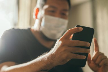 Koronavirüs karantinası kavramı. Tıbbi maskeli bir adam evde haber aramak için telefonu kullanıyor..