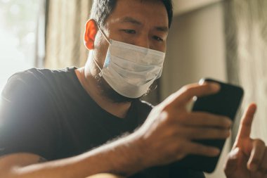 Koronavirüs karantinası kavramı. Tıbbi maskeli bir adam evde haber aramak için telefonu kullanıyor..