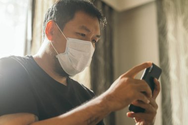 Koronavirüs karantinası kavramı. Tıbbi maskeli bir adam evde haber aramak için telefonu kullanıyor..