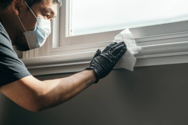 Coronavirus. Coronavirus 'u, Covid-19' u, gribi önlemek için koruyucu maskeli ve lateks eldivenli bir adam evi kağıt mendille temizliyor. Sprey şişesi. Virüs ve hastalık koruması. Beceriksiz..
