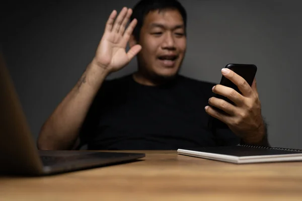 Asian man looking at cellphone with say hi on wooden table with laptop. Concept about work remotely.