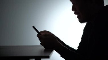 Siluet Asyalı adam akıllı telefon üzerinde çalışıyor. Beared Man cep telefonuyla evden çalışıyor. Ev 'de cep telefonuyla çalışan serbest çalışan blogcu adam. Bir adam bakıyor ve gülümsüyor.