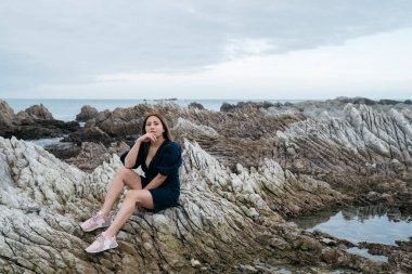 Deniz kenarındaki taşların üzerindeki Asyalı genç kadının portresi. Güzel kız kıyıda dinleniyor, Kaikoura, Yeni Zelanda 'da eğleniyor..