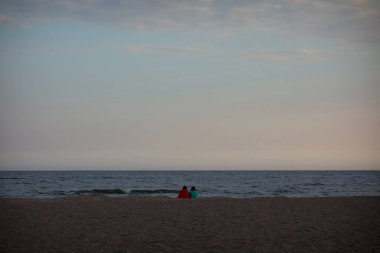 İki kişi sahilde oturur ve ufka bakar.