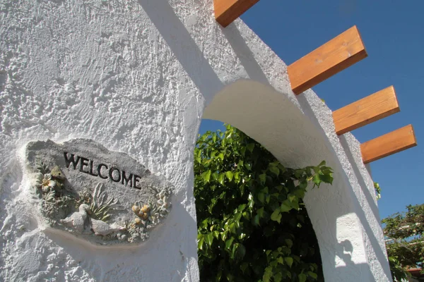 A welcome sign on a white wall in Greece, an arch with wooden projections.Sunny day.There are marine details on the plate.