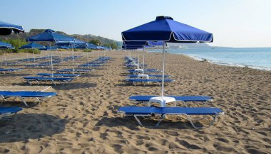 Rodos Faliraki kumsalında güneşli yataklı mavi şemsiyeler.