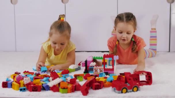Μικρά κορίτσια ξαπλώνουν στο χαλί και συναρμολογούν το σπίτι από πλαστικό λέγκο χτίζοντας τούβλα — Αρχείο Βίντεο