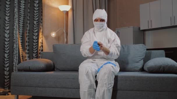 Мужчина в защитном костюме сидит дома на диване и надевает перчатки — стоковое видео