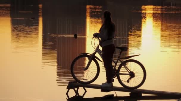 Силует жінки-велосипедистки, що стоїть на міському ставку з велосипедом на заході сонця — стокове відео