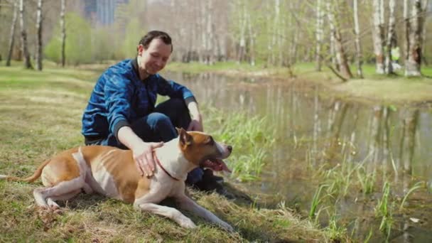 Ember ül a füvön a parkban a kutyájával Staffordshire terrier és simogatni őt — Stock videók