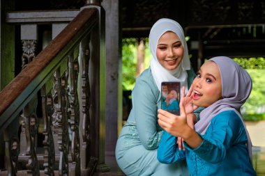 Geleneksel tesettür ve geleneksel kostüm giyen Müslüman Malezyalı kadınlar geleneksel ahşap evin terasında Aidilfitri kutlamaları sırasında selfie çekiyorlar. Hari Raya ve Müslüman kadınlar moda konsepti.
