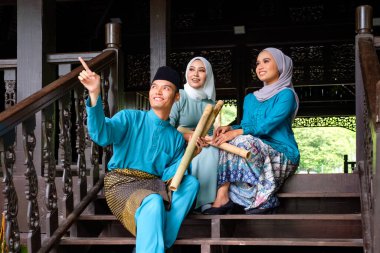 Geleneksel kostümlü üç Malaylı Müslüman 'dan oluşan bir grup geleneksel ahşap evin terasında Aidilfitri kutlaması sırasında mutlu bir konuşma yapıyorlar. Raya ve Müslüman moda konsepti.