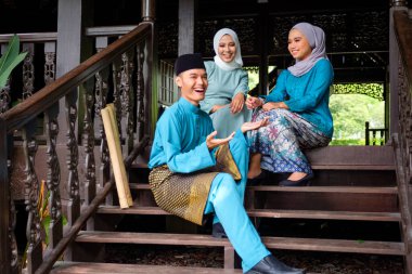 Geleneksel kostümlü üç Malaylı Müslüman 'dan oluşan bir grup geleneksel ahşap evin terasında Aidilfitri kutlaması sırasında mutlu bir konuşma yapıyorlar. Raya ve Müslüman moda konsepti.
