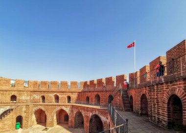 Alanya, Türkiye - 30 Eylül 2019: Gökyüzüne karşı güçlendirilmiş tuğla bir duvarın tepesinde Türk bayrağı. İki turist içeriden kuleyi ziyaret ediyor..