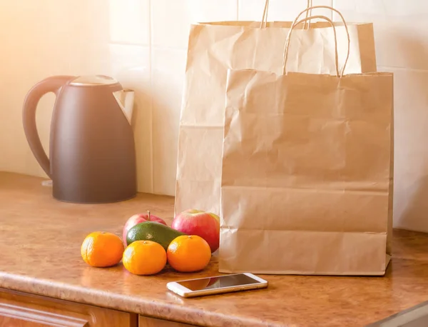 Paper bags, fresh vegetables and fruits next to a mobile phone on the table with kitchen utensils. Online shopping and contactless delivery concept.
