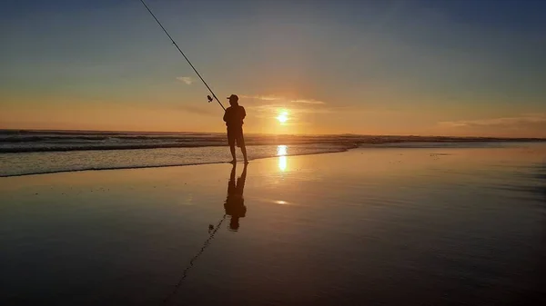 a single fishing man with a long rod at sunset time at Parangtritis Beach on wet and reflecting sand, Java, Indonesia