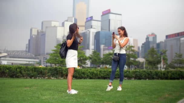 Две молодые женщины фотографируются в парке с зеленой травой и зданиями вокруг — стоковое видео