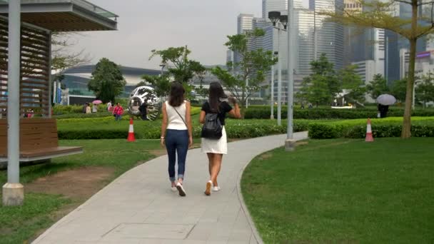 Две молодые женщины прогуливаются по парку с зелеными грассами и зданиями около 4k — стоковое видео