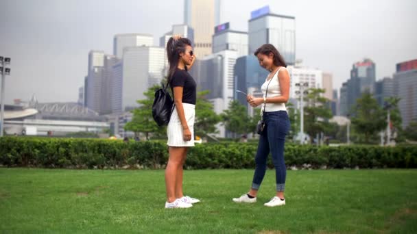 Dos mujeres jóvenes hablan en el parque con hierba verde y edificios alrededor de 4k — Vídeos de Stock