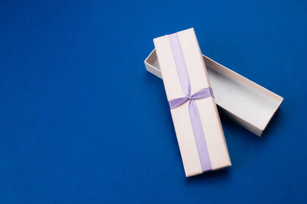 Small empty open gift box with purple ribbon flat lay on trendy blue background. Top view composition for birthday, valentine's day with copy space. Celebration concept. Template for web. Stock photo.