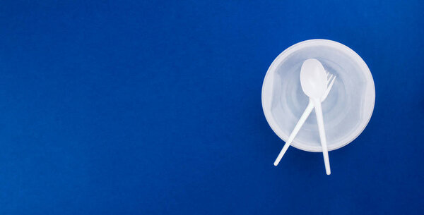 White plastic disposable tableware, utensil flat lay on trendy blue background. Contrast color. Plastic plate, bowl, spoon, fork. Top view copy space. Recycling concept. Banner template. Stock photo.
