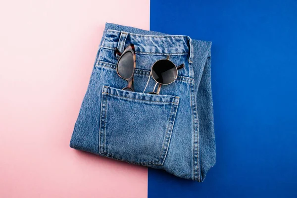 Nuevos vaqueros unisex azules y dos pares de gafas de sol planas yacen sobre un colorido fondo rosa y azul de moda. Todos los días estilo minimalista moda diseño vista superior. Plantilla para web, publicidad, redes sociales . — Foto de Stock