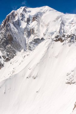 Karla kaplı kayalık dağ zirvesi yukarıdan esen rüzgarla açık mavi gökyüzüne doğru...