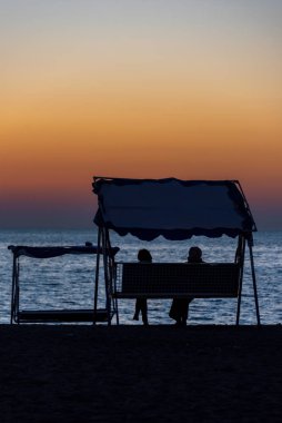 Balkonda salıncakta sallanan iki kişinin silueti sahilde gün batımını seyrediyor.
