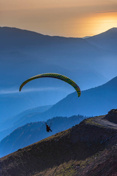 Paraglider flies by a mountain dirt road with a series of valleys, clouds, mountains and the golden sunset sea in the background