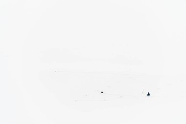 Tek başıma. Beyaz arka planda elinde çengelle oturan yalnız bir buzdan balıkçı, yukarıdan el makasıyla çekilmiş yalnız bir buzdan balıkçı. Ayak izleri buzu ve sırt çantasını kaplayan karda zar zor görülebiliyor..