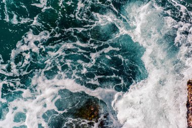 Wave crashes on the rocks creating a swirl of white foam on the azure blue water of the sea