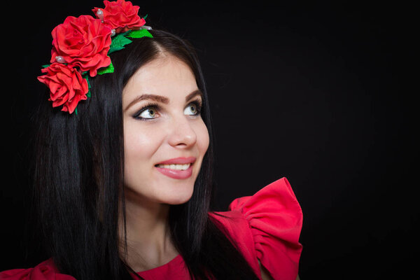 Woman with long black hair and a wreath with red flowers on a dark background smiles and looks away. Close-up. Space for text