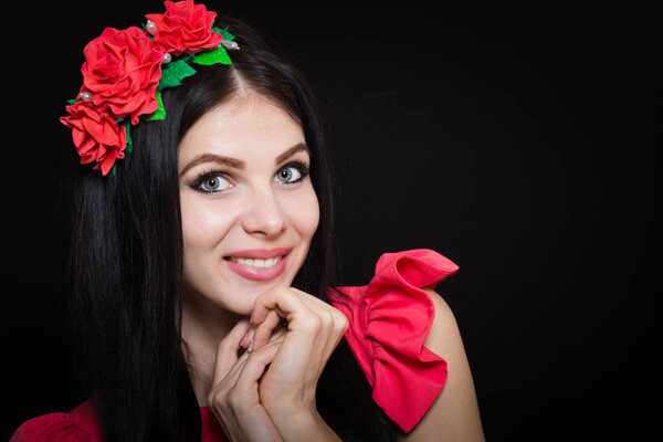 Woman with long black hair and wreath with red flowers on a dark background smiles. Close-up. Space for text