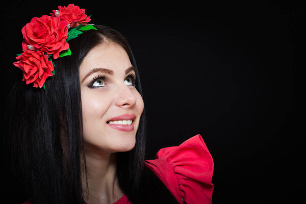 Woman with long black hair and a wreath with red flowers on a dark background smiles and looks away. Close-up. Space for text