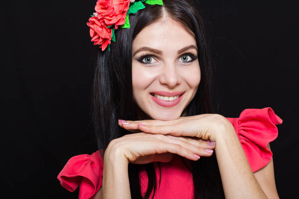 Woman with long black hair and wreath with red flowers on a dark background smiles. Close-up. Space for text
