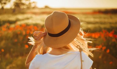 Haşhaş tarlasında sarışın bir kadın şapkasını çeker ve güneşe doğru bakar. Yakın plan.
