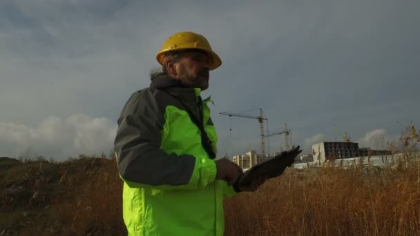 Mature male worker foreman in helmet with a tablet on background of construction — Stock Video