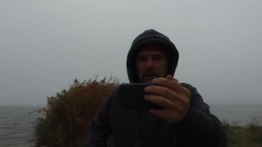 Akıllı telefondan görüntülü konuşma. Sakallı, olgun bir adam bir sonbahar nehrinin kıyısında uzanmış elinde telefonu tutuyor.