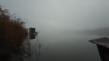 Şiddetli sisli yaz nehri manzarası, balıkçı evi ve sazlıklar suya yansıyor.