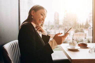 Ofis kıyafetleri içindeki güzel bir kız ve elinde inanılmaz bir gülümsemeyle kafede oturmuş güneşli bir günde masada kek yiyor.