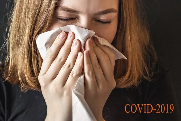 A sick young woman with a wrinkled nose, blowing her nose and wrinkled, caught a cold, sneezes in tissue, coughs, and has the flu. On a black background the inscription \