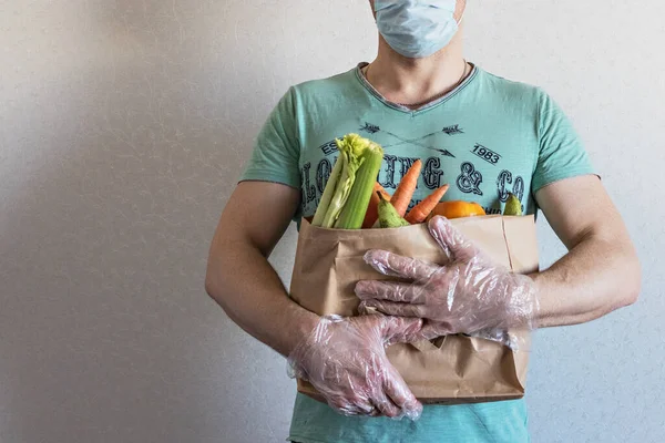 Bir adam elinde alışveriş poşeti olan bir eco kağıdı tutuyor. Eve kurye ile yemek teslimatı. Eldivenli eller. Coronavirus, COVID 19