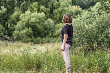 Genç bir kadın arkası ormana dönük duruyor. Yaz zamanı..