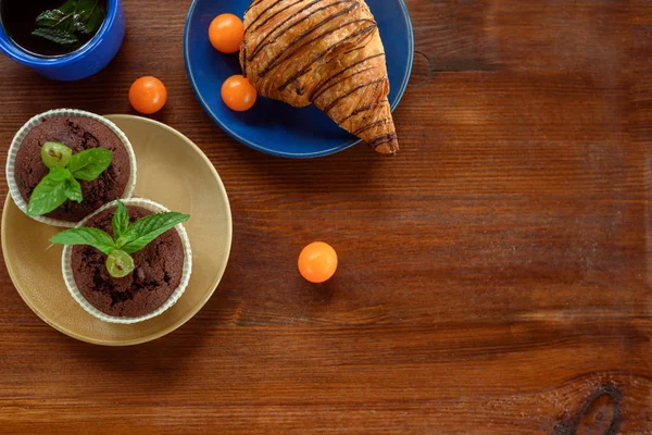 Sabah kahvaltısı: mavi bardak çay, çikolata kruvasan, kekler ve physalis ahşap bir masa üzerinde. Düz kompozisyon