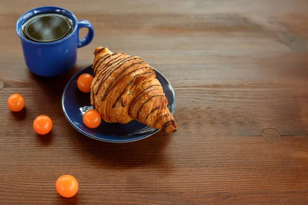 Frühstück am Morgen: Blaue Tasse Tee, Schokocroissant und Physalis auf einem Holztisch — Stockfoto