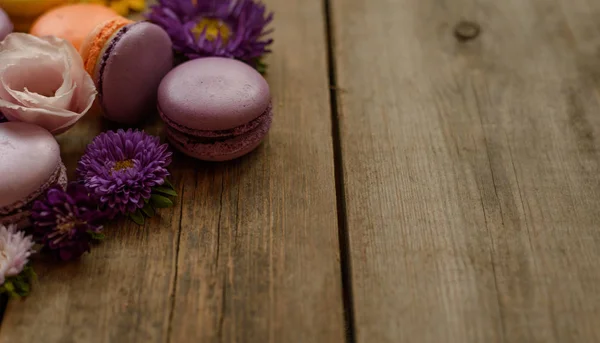 Фиолетовый и желтый макароны и цветы на фоне деревянного стола. Цветной французский десерт со свежими цветами — стоковое фото