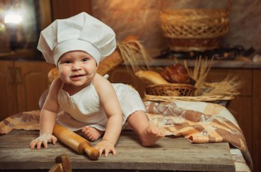 Mutlu küçük çocuk aşçı kostüm ile lezzetli rulo, ekmeği ve ekmek sepetleri arasında masadaki oturan mutfak at. Kopya alanı
